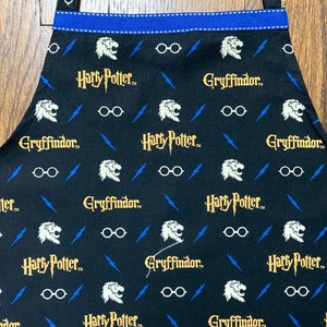 May include: Black kitchen apron with a repeating pattern of Harry Potter and Gryffindor logos, lightning bolts, and glasses. The apron has a blue trim.