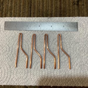 May include: Four identical bent copper wire components, each featuring a narrow U-shaped top and an angled lower section. A metal ruler is positioned above the pieces, showing each component is approximately 2.5 inches in total height.