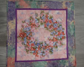 May include: A quilted floral table runner with a central square of pink fabric featuring a wreath of red, blue, and white flowers. The square is framed by a purple border and surrounded by a tie-dye patterned fabric.