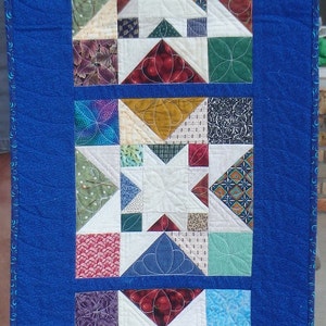 Puede incluir: Un camino de mesa acolchado azul con un patr&oacute;n repetido de estrellas blancas y cuadrados de tela de colores. El camino de mesa est&aacute; formado por cinco filas de estrellas y cuadrados.