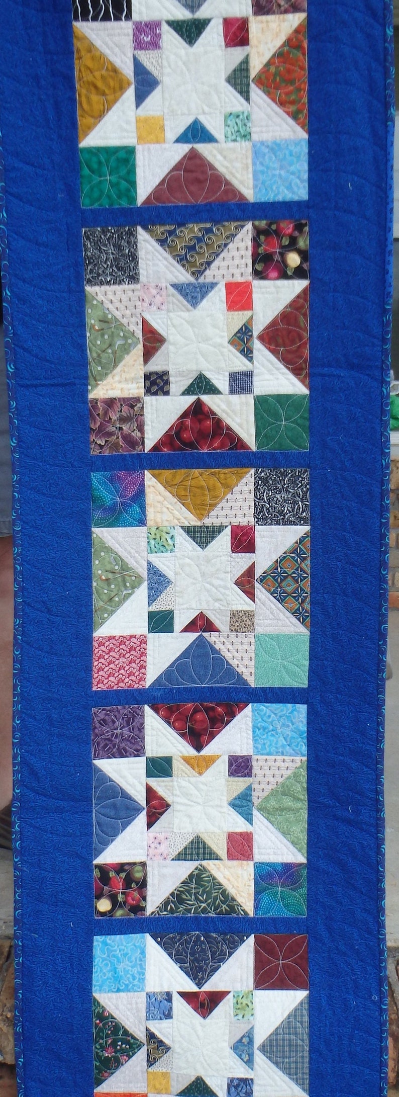 Puede incluir: Un camino de mesa acolchado azul con un patr&oacute;n repetido de estrellas blancas y cuadrados de tela de colores. El camino de mesa est&aacute; formado por cinco filas de estrellas y cuadrados.