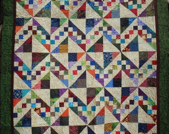 Op de afbeelding: Een patchwork quilt met een herhalend geometrisch patroon van crèmekleurige driehoeken afgewisseld met kleine, veelkleurige vierkantjes in tinten rood, blauw, groen, paars en oranje, afgewerkt met een donkerbruine rand en een brede groene boord.