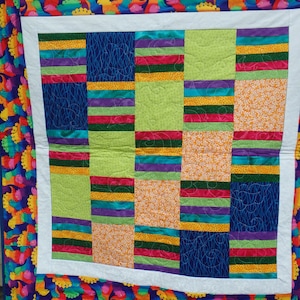 Può includere: Una trapunta colorata con un bordo a stampa di dinosauro. La trapunta è composta da una griglia di quadrati con diversi motivi e colori, tra cui verde, blu, arancione, giallo, rosso e viola.