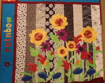 May include: A colourful quilt with the words "in my garden" at the top and "a rainbow" on the left side. The quilt features a variety of flowers, including sunflowers, daisies, and tulips, in a black and white patterned background.