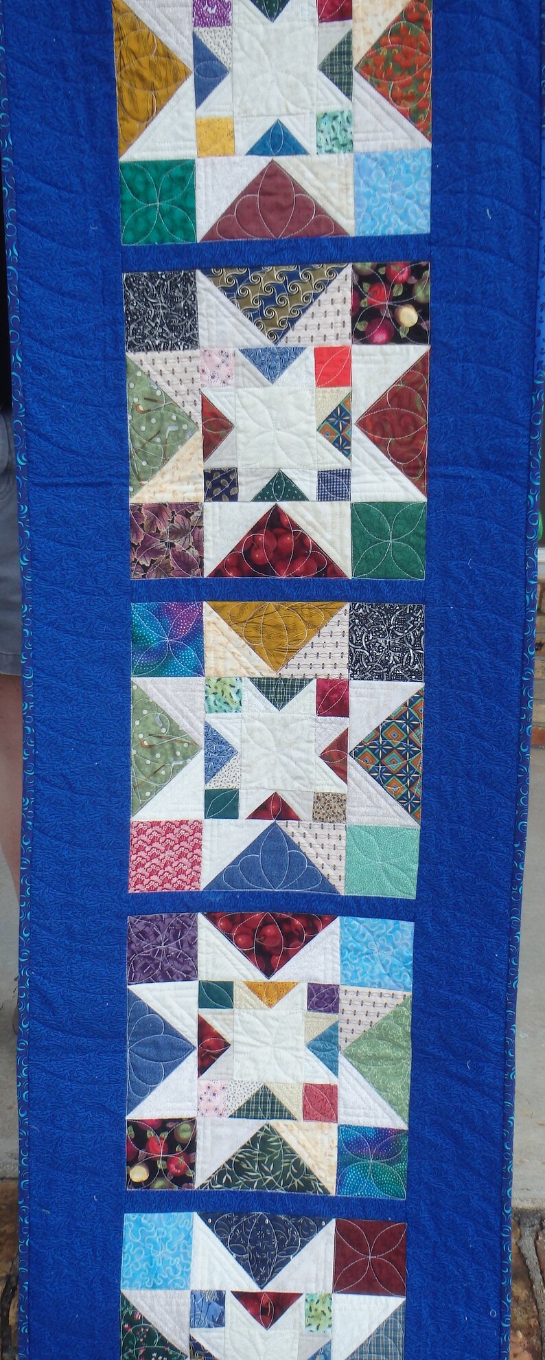 Puede incluir: Un camino de mesa acolchado azul con un dise&ntilde;o de patchwork que presenta varios patrones y colores. El camino de mesa est&aacute; formado por varios cuadrados con diferentes telas, incluyendo dise&ntilde;os florales, geom&eacute;tricos y abstractos. El camino de mesa est&aacute; bordeado con un ribete azul.