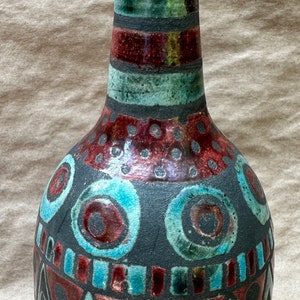 May include: A ceramic vase with a narrow neck and bulbous body. The vase features a dark base with turquoise, red, and black geometric patterns. The neck has horizontal bands of red, green, and black. A decorative piece.