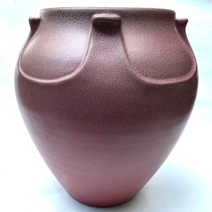 May include: A mauve-colored ceramic vase with a textured surface and a rounded body. The vase features two curved handles on each side of the opening. The color transitions from a darker shade at the top to a lighter shade at the base.