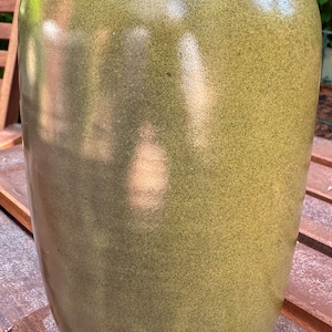 May include: A tall, olive green ceramic vase with a speckled texture. The vase has a rounded body and a slightly flared rim. The surface reflects light, creating highlights. This decorative vase is suitable for home decor.