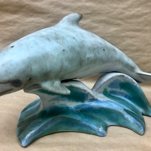 Peut inclure: Une sculpture en céramique d'un dauphin sautant hors de l'eau. Le dauphin est peint dans des tons de bleu et de blanc, et l'eau est peinte dans des tons de bleu et de vert.