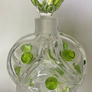 May include: Clear glass perfume bottle with a round body and a decorative stopper. The bottle is adorned with green glass accents. The stopper has a swirl design with green accents. The bottle is empty.