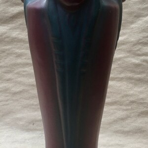 Könnte beinhalten: Eine dunkelgrüne und burgunderrote Keramikvase mit einem indianischen Gesichtsdesign. Die Vase hat einen breiten Boden und einen schmalen Hals.