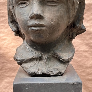 May include: A gray stone bust sculpture of a child with short hair. The bust is on a black square base.