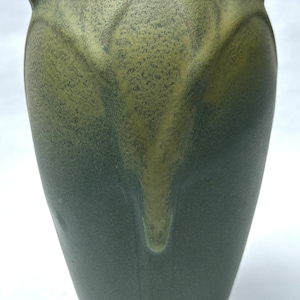 May include: A tall, green ceramic vase with a textured surface and a unique, leaf-like design. The vase has a wide base that tapers upwards to a narrower neck with a flared rim. The color gradient transitions from a darker green at the base to a lighter, olive green at the top.