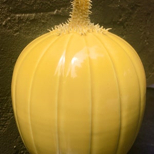 Puede incluir: Una calabaza de cerámica amarilla con un tallo espinoso. La calabaza tiene un acabado brillante y está decorada con crestas verticales.