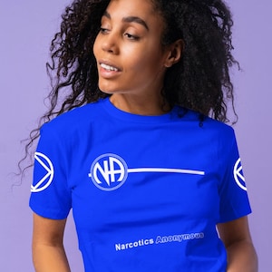 May include: A woman wearing a blue t-shirt with a white circle containing the letters "NA" and the text "Narcotics Anonymous".