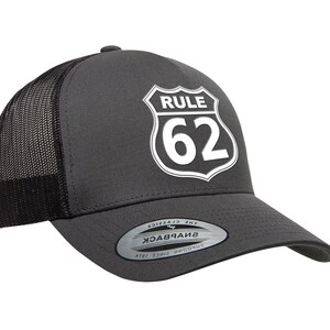 May include: Charcoal gray trucker hat with a black mesh back and a curved brim. The front features a white graphic of a road sign with the text "RULE 62". The hat has a snapback closure.