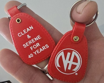 Puede incluir: Dos llaveros rojos con anillas plateadas. Un llavero tiene el texto blanco "CLEAN & SERENE FOR 40 YEARS". El otro llavero tiene un logotipo blanco con las letras "NA" dentro de un círculo.