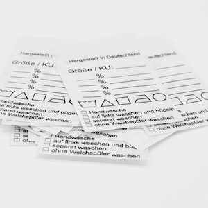 Könnte beinhalten: Ein Stapel weißer Pflegeetiketten für Kleidung mit schwarzem Text und Symbolen. Die Etiketten enthalten Anweisungen zum Handwaschen, Bügeln und Trocknen. Die Etiketten haben auch Platz für die Größe und andere Informationen.