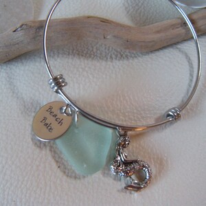 May include: A silver bangle bracelet with a charm that says "Beach Babe" and a sea glass charm. The bracelet also has a mermaid charm.