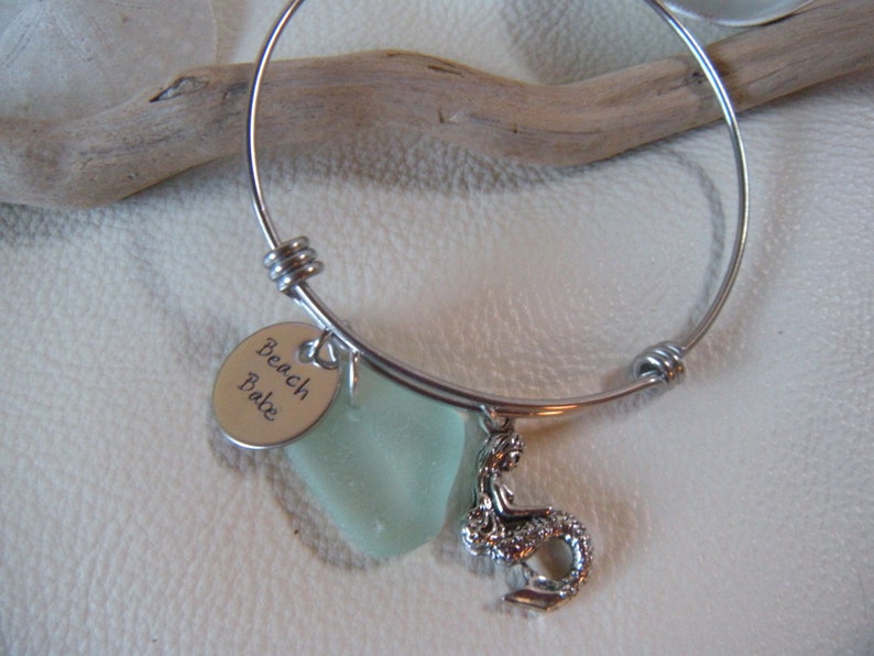 May include: A silver bangle bracelet with a charm that says "Beach Babe" and a sea glass charm. The bracelet also has a mermaid charm.