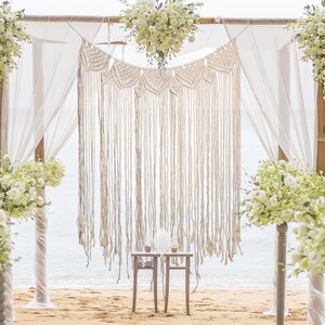 May include: A white macrame wedding backdrop with a delicate fringe, hanging from a wooden archway decorated with white flowers. Two small wooden tables with white tops are placed in front of the backdrop.