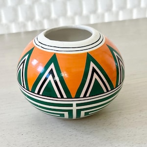 May include: A small, round, orange and green ceramic pot with a geometric design. The pot is hand-painted with a white rim and a black outline.