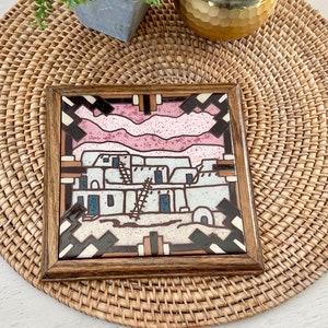 Könnte beinhalten: Quadratisches, gerahmtes Kunstwerk mit einem südwestlichen Design. Das Kunstwerk zeigt eine Dorfszene mit rosa und weißen Elementen, vor einem braunen und schwarzen geometrischen Rahmen. Der Rahmen ist aus Holz, und das Kunstwerk wird auf einem gewebten Platzset präsentiert.