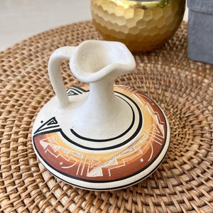 May include: A small, white ceramic pitcher with a brown and yellow geometric design. The pitcher has a rounded body and a small, curved handle. It is sitting on a woven wicker placemat.
