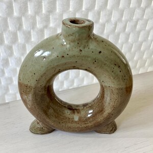 May include: A ceramic vase with a donut shape and a green and brown glaze. The vase has three small feet.