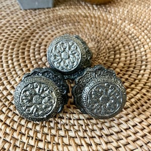May include: Three black metal drawer pulls with a decorative floral design. The pulls have a distressed finish and are arranged on a woven wicker surface.