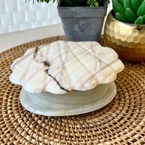May include: A white and brown marble trinket box with a scalloped lid. The box is sitting on a woven wicker mat.