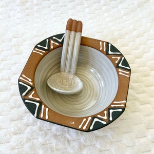 May include: A white ceramic bowl with a brown rim and black and white geometric design. The bowl is paired with a matching white ceramic spoon.