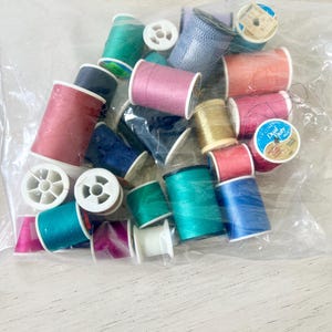 May include: A clear plastic bag filled with colorful spools of thread. The spools are various colors, including pink, teal, blue, and gold. Some spools are labeled. The thread is for sewing and crafting.