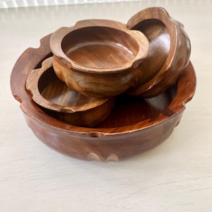 Pode incluir: Conjunto de quatro tigelas de madeira, aninhadas umas nas outras. As tigelas são de madeira castanho escura e têm um acabamento liso e polido. A tigela maior tem uma borda festonada.