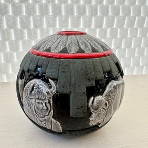 May include: A black ceramic vase with a red rim and a white design of Native American figures and feathers.