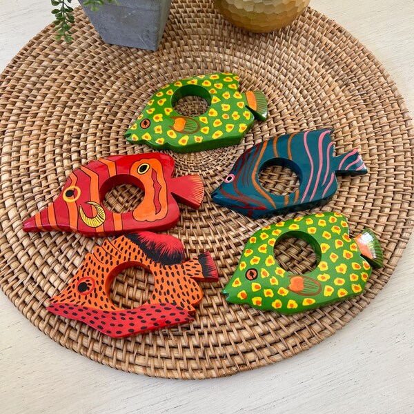 Puede incluir: Cinco servilleteros de madera pintados a mano con forma de pez, en colores vibrantes. Los peces están pintados con intrincados patrones en rojo, naranja, verde y azul. Están dispuestos sobre un salvamanteles tejido, perfecto para una mesa de temática náutica o tropical.