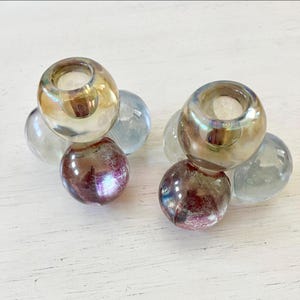 May include: Two glass candle holders. Each holder has a spherical base in shades of purple and clear, supporting a larger, iridescent, spherical top. Clear glass spheres surround the base of each holder. The holders are on a white surface.