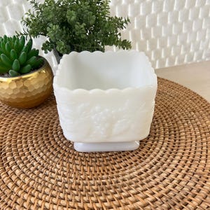 May include: A white, square, milk glass dish with a scalloped rim and embossed floral design. The dish sits on a small, rectangular base. The dish is displayed on a woven, round placemat.