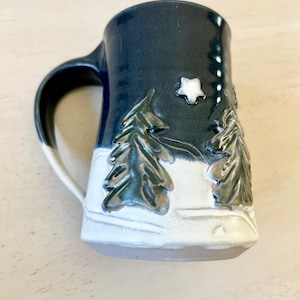 May include: A ceramic mug with a dark blue exterior and a white interior. The mug features a winter scene with embossed evergreen trees and a star. The handle is black and white.