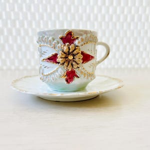 May include: A decorative teacup and saucer set. The cup and saucer are white with gold trim. The cup features a red and gold floral design. The set is likely vintage or antique.