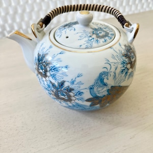 May include: A white porcelain teapot with a blue and gold floral design featuring a peacock. The teapot has a brown woven handle and a lid with a small hole.