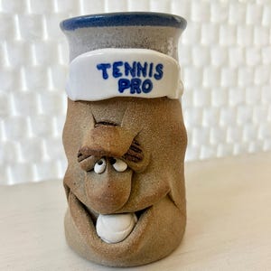 May include: A whimsical ceramic vase with a face design. The vase is primarily tan with a white band that reads "TENNIS PRO" in blue. The top rim is blue, and the face has exaggerated features, including a large white tooth.