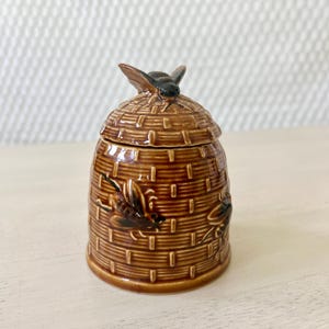 May include: A brown ceramic honey pot shaped like a beehive, with a bee figurine on top. The pot has a woven texture and bee illustrations on the sides. The lid fits snugly, and the pot is designed for storing honey.