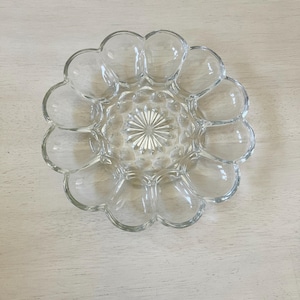 May include: Clear glass egg serving dish with a flower-like design. The dish has a central starburst pattern and individual egg-shaped compartments around the perimeter. The dish is made of clear glass and is designed to hold a dozen eggs.