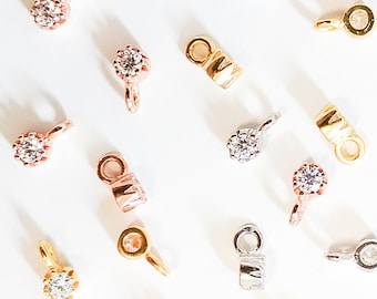 May include: Assortment of silver, gold, and rose gold tone metal charms with clear rhinestone accents. The charms feature different shapes and designs, including round, square, and rectangular.