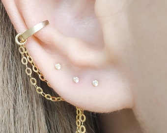 Può includere: Un orecchino a cerchio dorato con una catena pendente e tre piccoli diamanti nel lobo dell'orecchio.