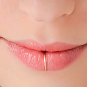 Pode incluir: Um close-up de um lábio inferior com um pequeno piercing de anel de ouro. O anel é fino e delicado, e está colocado no centro do lábio.