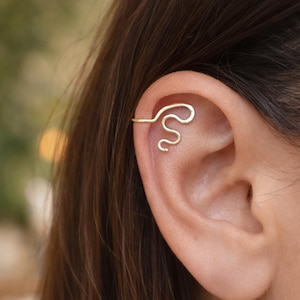 May include: A gold-colored ear cuff in a snake-like design. The jewelry curves along the outer ear, creating a unique and stylish accessory. The cuff is made of a thin, wire-like material, and is worn on the upper ear.