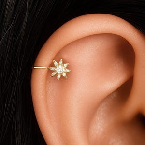 Puede incluir: Un pendiente de oreja dorado con un diseño de estrella, adornado con pequeñas piedras preciosas transparentes. También se ven tres pendientes pequeños y redondos con piedras preciosas en la oreja. La joya se muestra sobre una oreja y cabello oscuro.