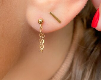 Puede incluir: Primer plano de la oreja de una persona con tres pendientes de oro. Los pendientes son una pequeña bola con una cadena, una pequeña barra y un aro.